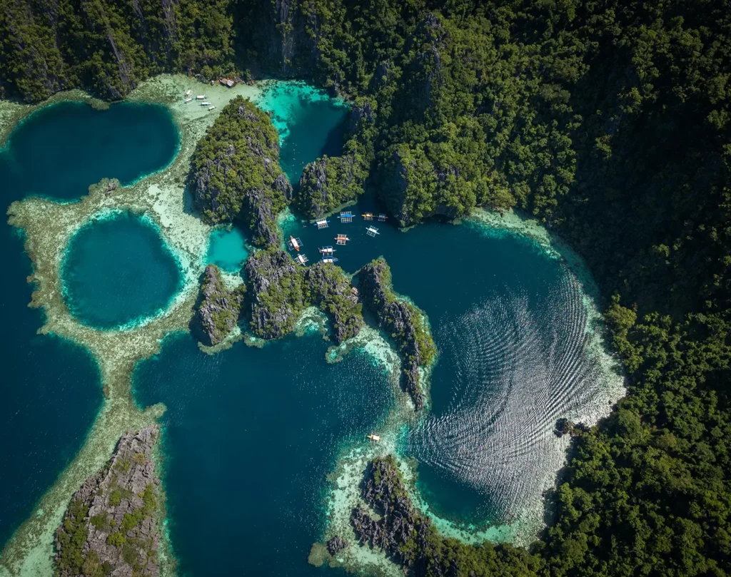 Aerial top-down view of vibrant turquoise lagoons, coral reefs, and karst limestone formations in El Nido, Palawan, dotted with outrigger boats.