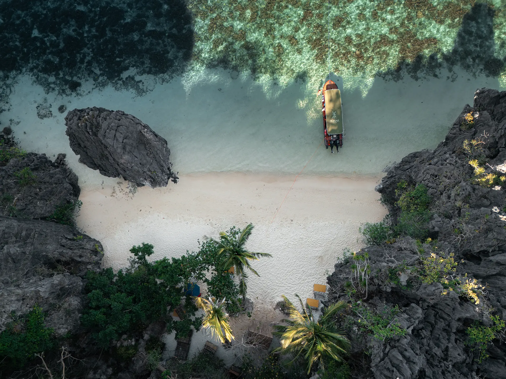 Top-down drone view of the Yohak speedboat anchored at a secluded white sand beach between dark limestone cliffs in El Nido.