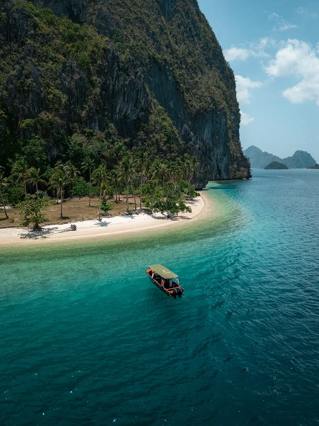 Wide drone shot of the Yohak tour boat navigating the tropical seascape of El Nido, sitting below massive limestone karst cliffs.