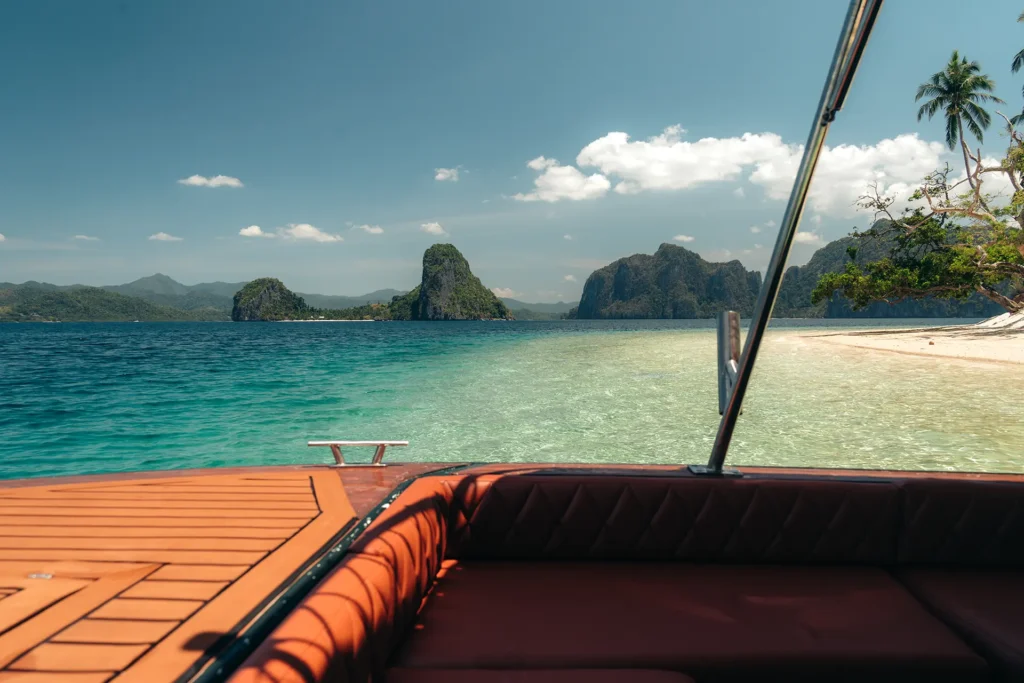 First-person view from the wooden deck and leather seating of a speedboat looking out over clear water toward a white sand beach and limestone karst islands in El Nido.