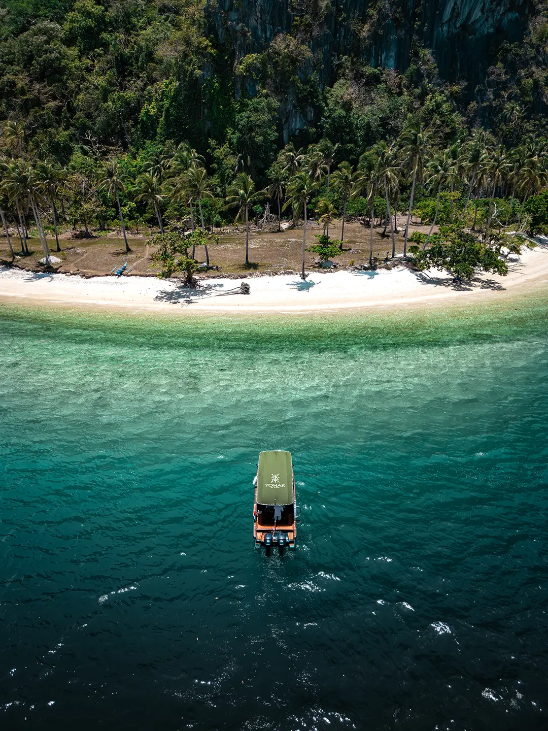 Aerial view of the Yohak speedboat anchored in clear water directly in front of a white sand beach and palm trees in El Nido.
