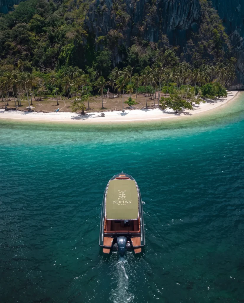 Aerial view of a Yohak El Nido speedboat approaching a tropical beach with palm trees and limestone cliffs in Palawan.