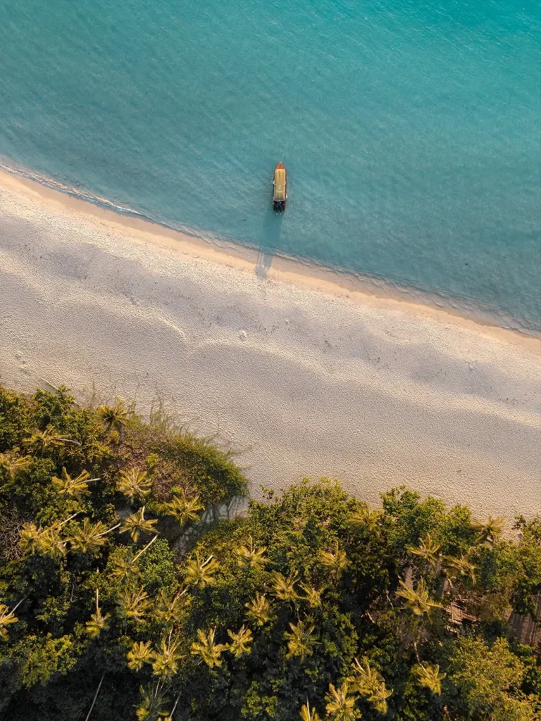 Aerial view of a pristine white sand beach with crystal-clear turquoise water, a single outrigger boat anchored offshore, and lush green coastal vegetation framing the bottom edge.